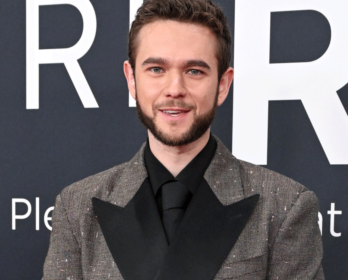 A man with short brown hair and a beard wears a dark glittery suit and black shirt and tie, standing in front of a dark background with large white letters.