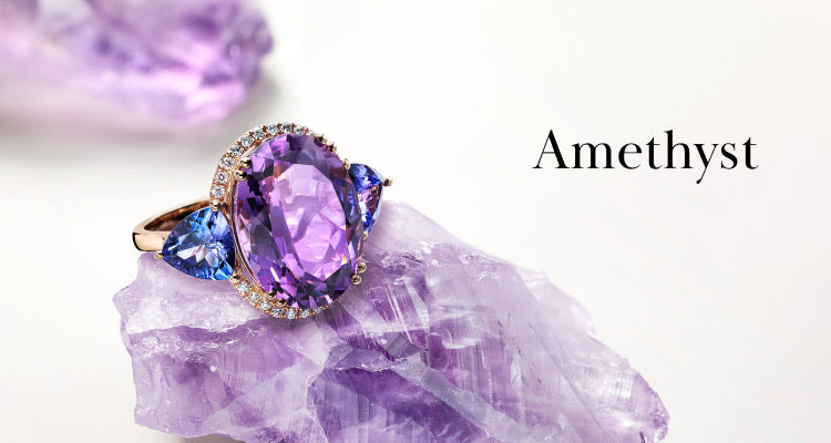 A gold ring with a large oval amethyst gemstone, flanked by two blue stones and small diamonds, rests on a piece of raw amethyst. The word Amethyst appears on the right against a white background.