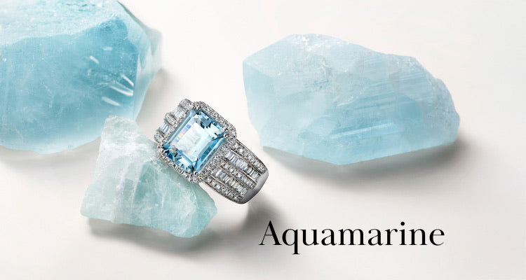A silver ring with a large, rectangular aquamarine gemstone surrounded by diamonds rests on a light blue aquamarine crystal, with two more crystals nearby and the word Aquamarine written on the white background.
