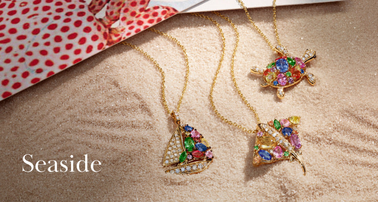 Three gold necklaces with colorful gemstone pendants—a sailboat, a turtle, and a butterfly—are displayed on sandy textured surface with a magazine in the corner. Text reads Seaside.