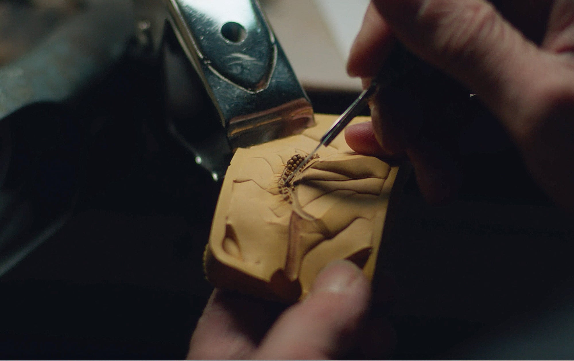 A close-up of hands using a small tool to carve detailed patterns into a piece of tan-colored clay or sculpting material, held steady by a metal clamp.