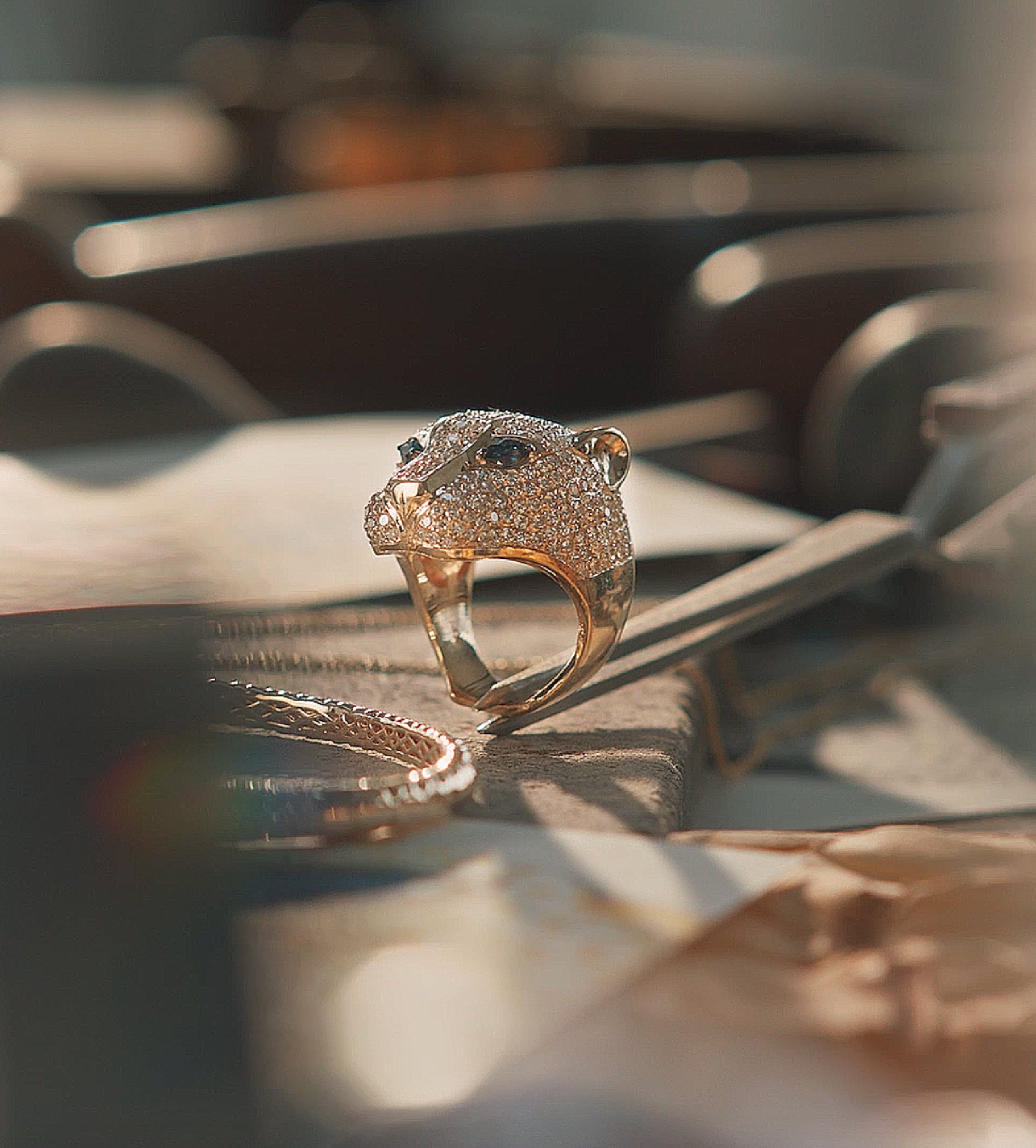 A gold ring shaped like a panthers head, encrusted with diamonds and featuring dark gemstone eyes, sits on a soft beige surface with blurred objects in the background.