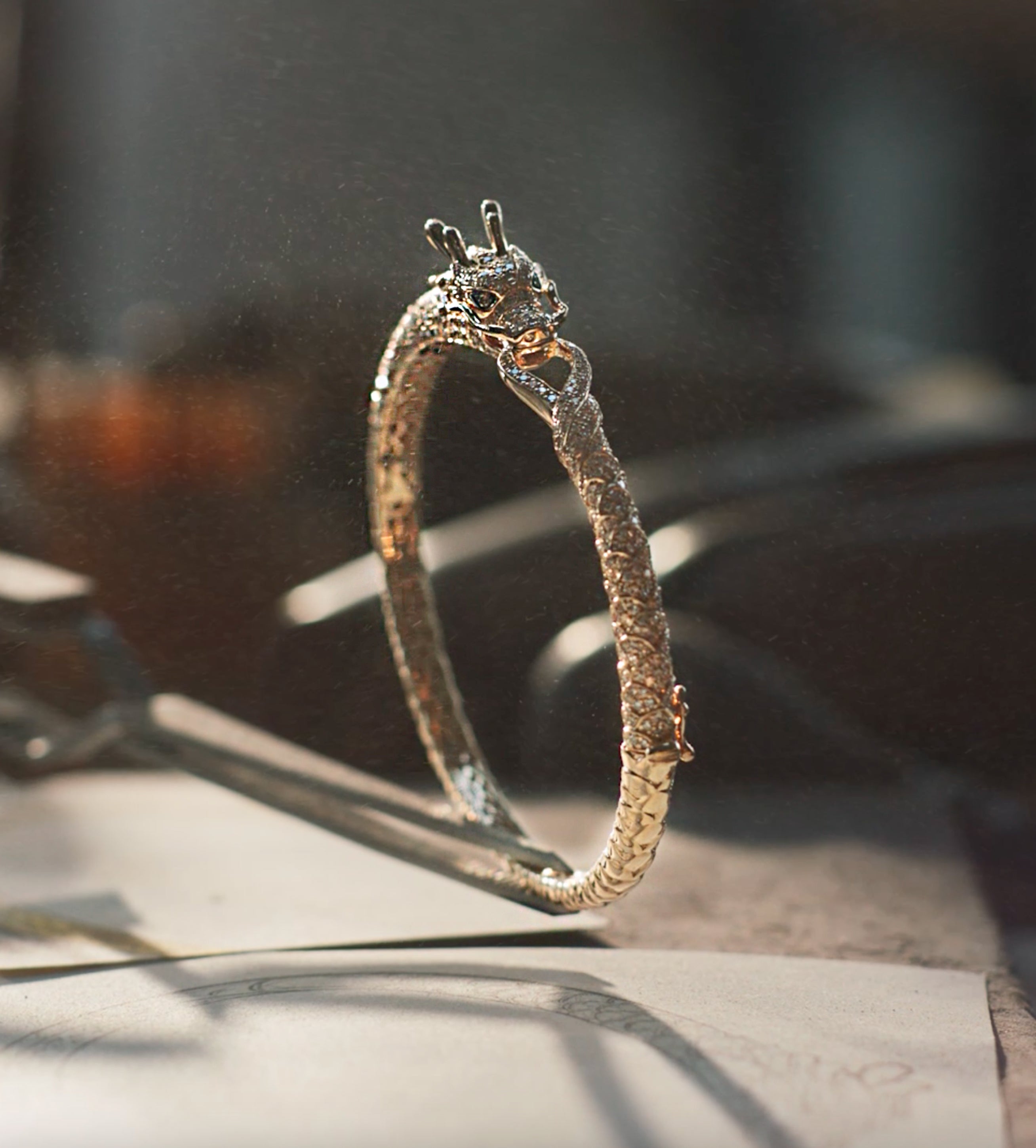 A detailed silver bracelet shaped like a dragon, with the dragon’s head and tail meeting at the front, is displayed upright on a surface with soft, warm lighting and a blurred background.