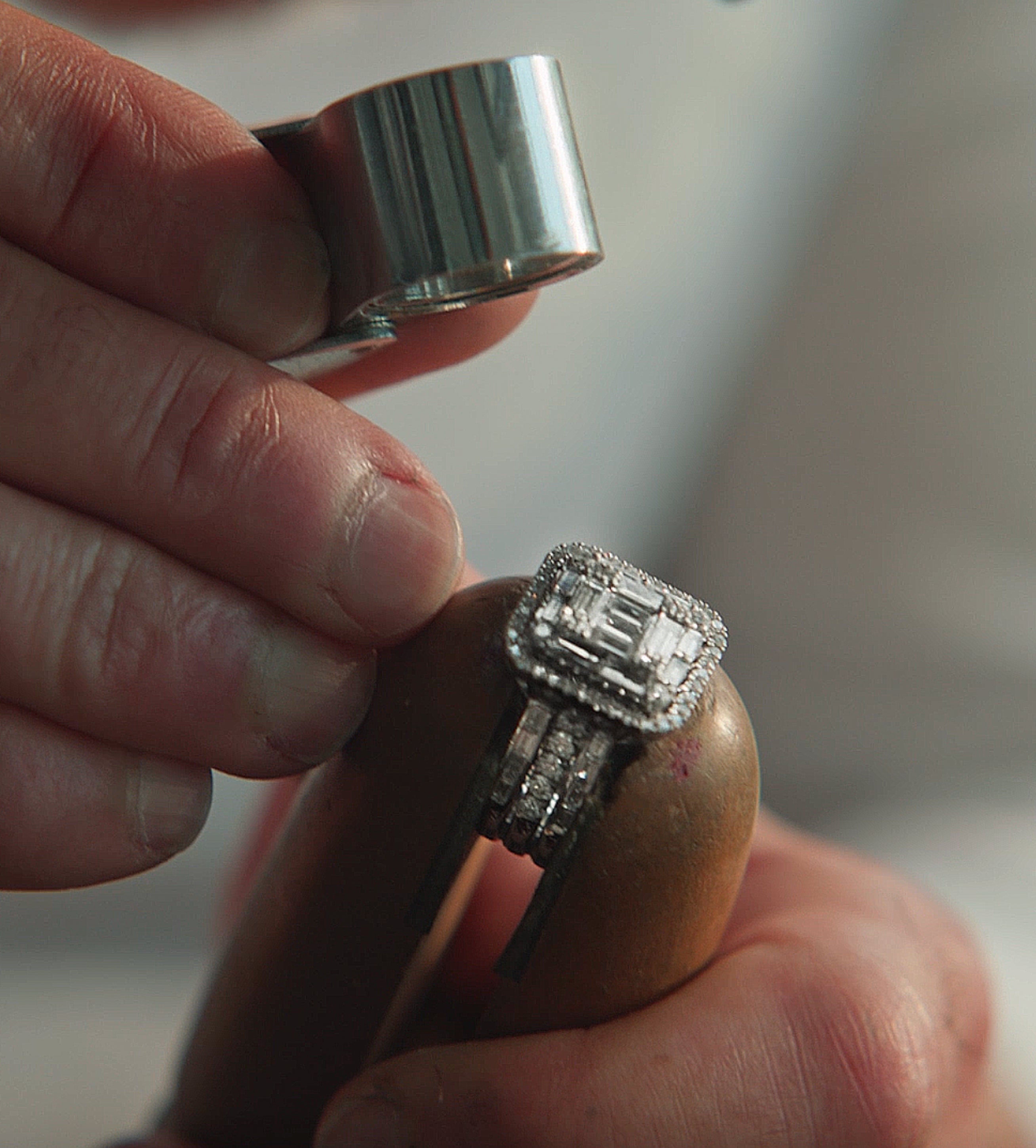 A person examines a diamond ring with a jeweler’s loupe, holding the ring securely in a wooden clamp. The ring features a large central stone surrounded by smaller diamonds.
