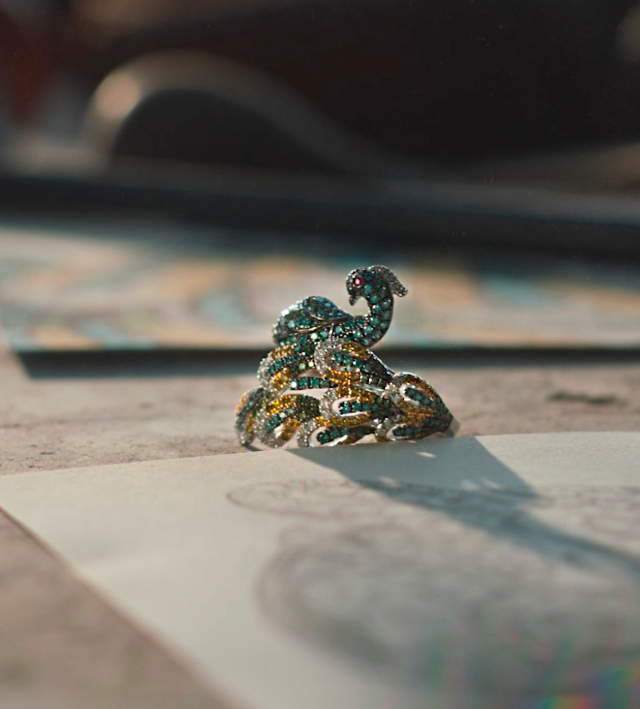 A jeweled ring shaped like a coiled snake with sparkling green, yellow, and white stones sits on a table, casting a shadow over a piece of paper with a sketch.