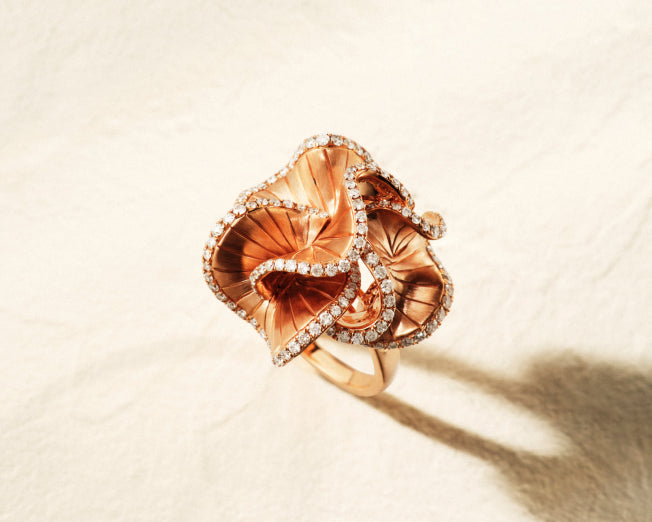 A gold ring designed in the shape of a blooming flower, featuring intricate petals bordered with small sparkling diamonds, resting on a light, textured surface.