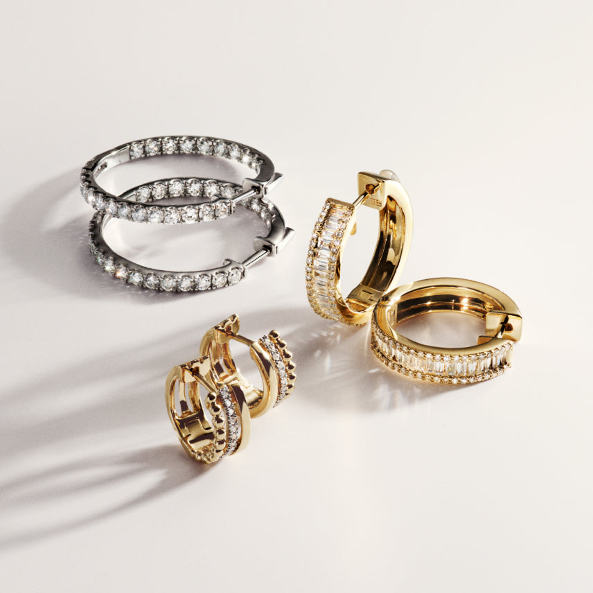 Three pairs of hoop earrings—one silver and two gold—are displayed on a white surface. Each pair features rows of sparkling clear stones set along the outer and inner edges. Shadows are cast beside the earrings.