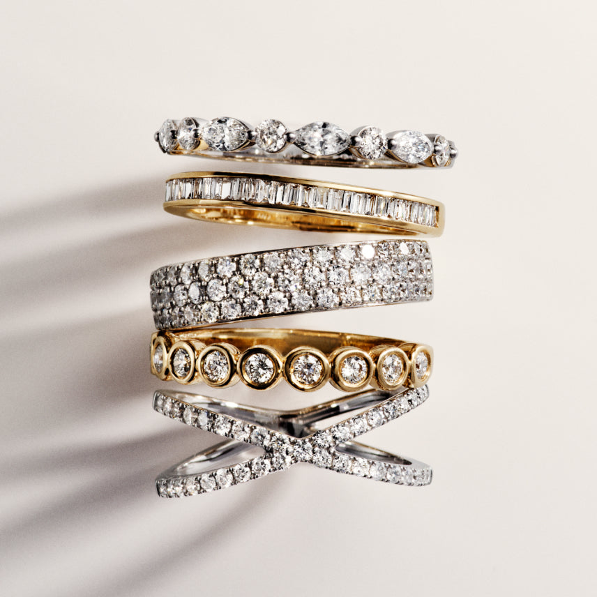 A stack of six gold and silver rings, each adorned with various arrangements of sparkling diamonds, displayed against a light background.