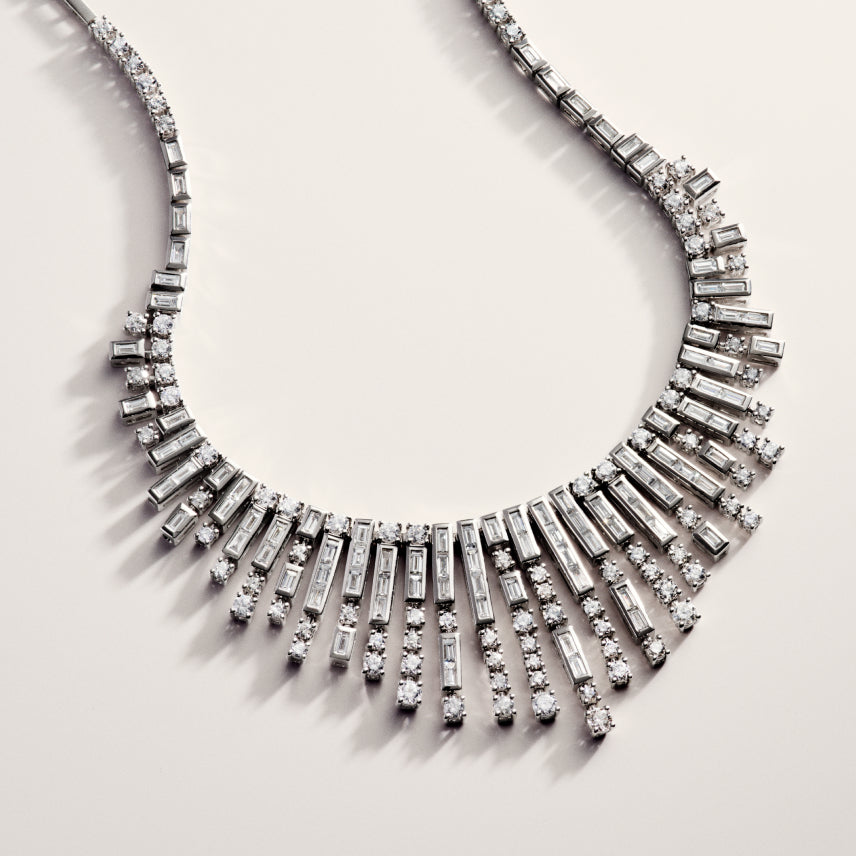 A silver necklace with rectangular and round clear gemstones arranged in a fan-like, graduated pattern, creating a sparkling, elegant design on a light background.