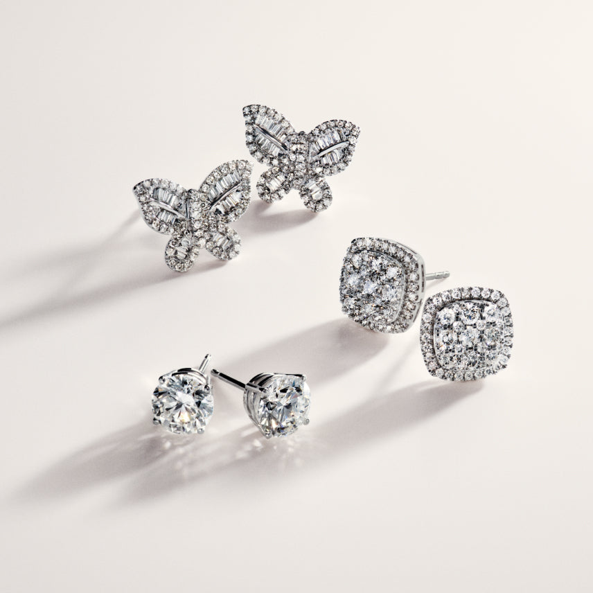 Three pairs of diamond stud earrings are displayed: butterfly-shaped, square-shaped with a halo setting, and round solitaire studs, all on a light, neutral background.