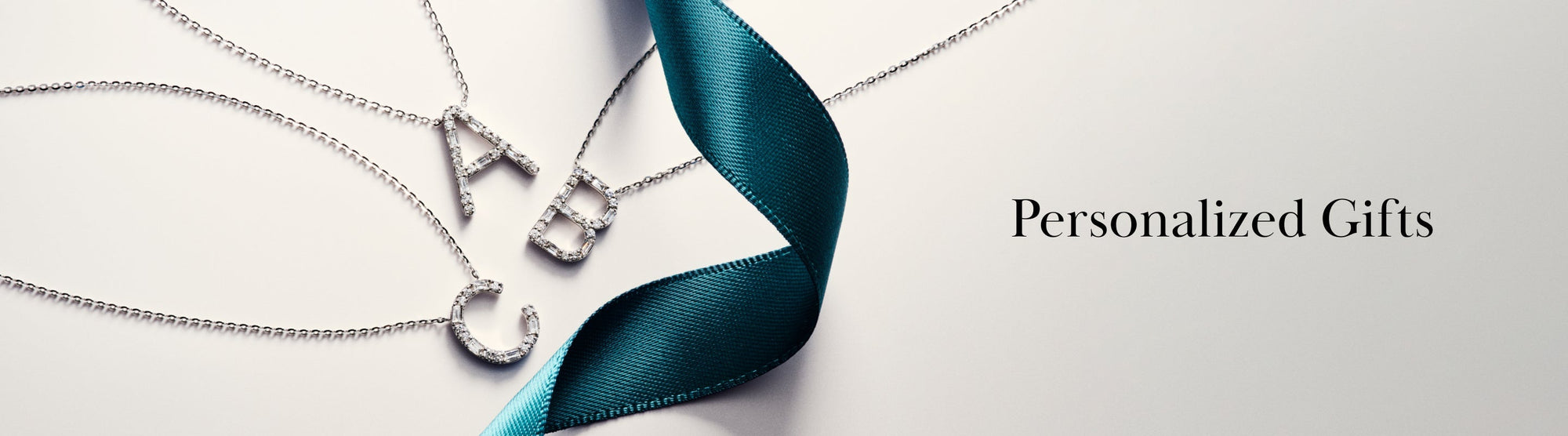 Three silver necklaces with letter pendants A, B, and C lay on a pale surface beside a teal ribbon, with the text Personalized Gifts written on the right.