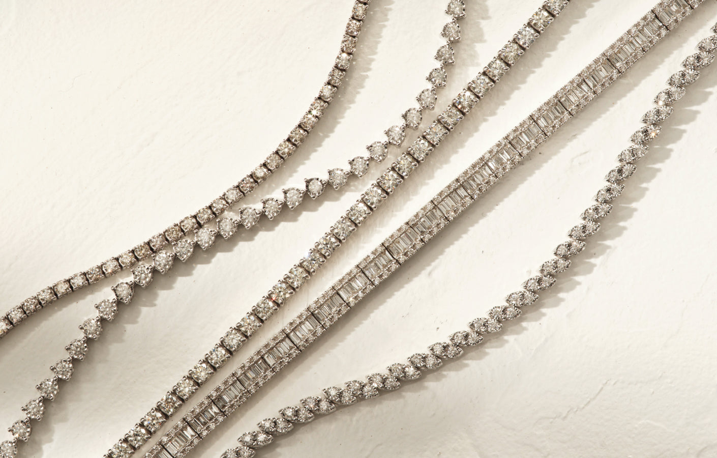 Several elegant diamond tennis bracelets with different stone shapes and settings are arranged diagonally on a light textured surface, sparkling under soft lighting.