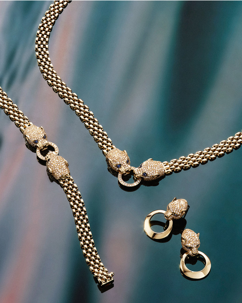 Gold jewelry set consisting of a necklace, bracelet, and earrings featuring textured links and encrusted panther head clasps, photographed on a blue and pink gradient background.