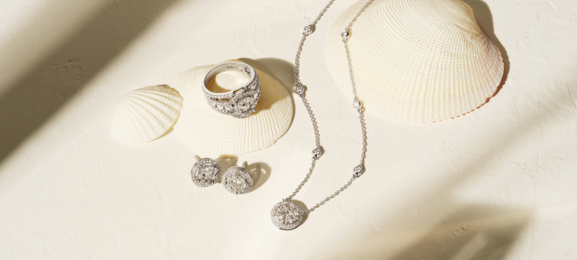 Silver jewelry set, including a ring, pair of earrings, and a necklace with round, sparkling stones, displayed on a light surface with seashells and soft shadows.