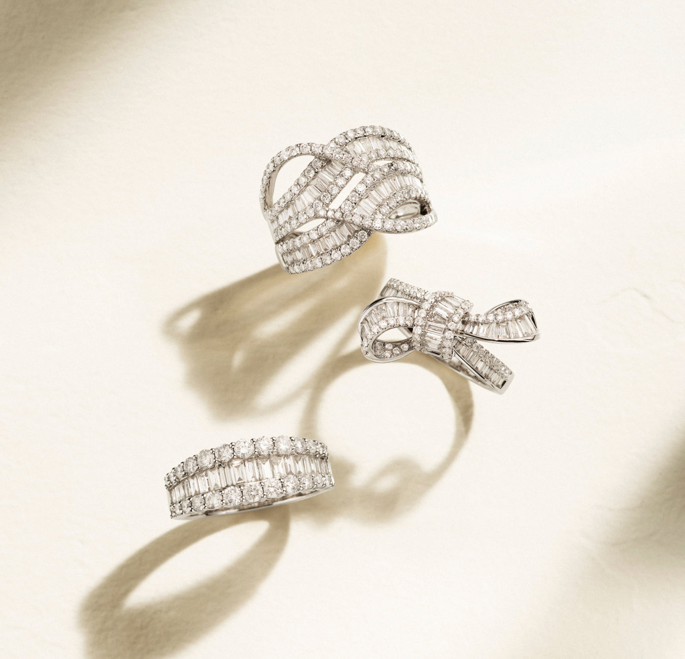 Three elegant diamond rings with intricate, sparkling designs are displayed on a light, textured surface, casting soft shadows.