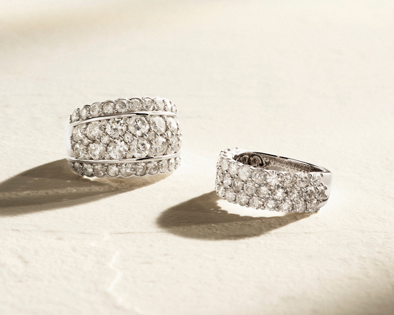 Two silver rings adorned with rows of sparkling white diamonds rest on a textured, light-colored surface, casting soft shadows in natural light.