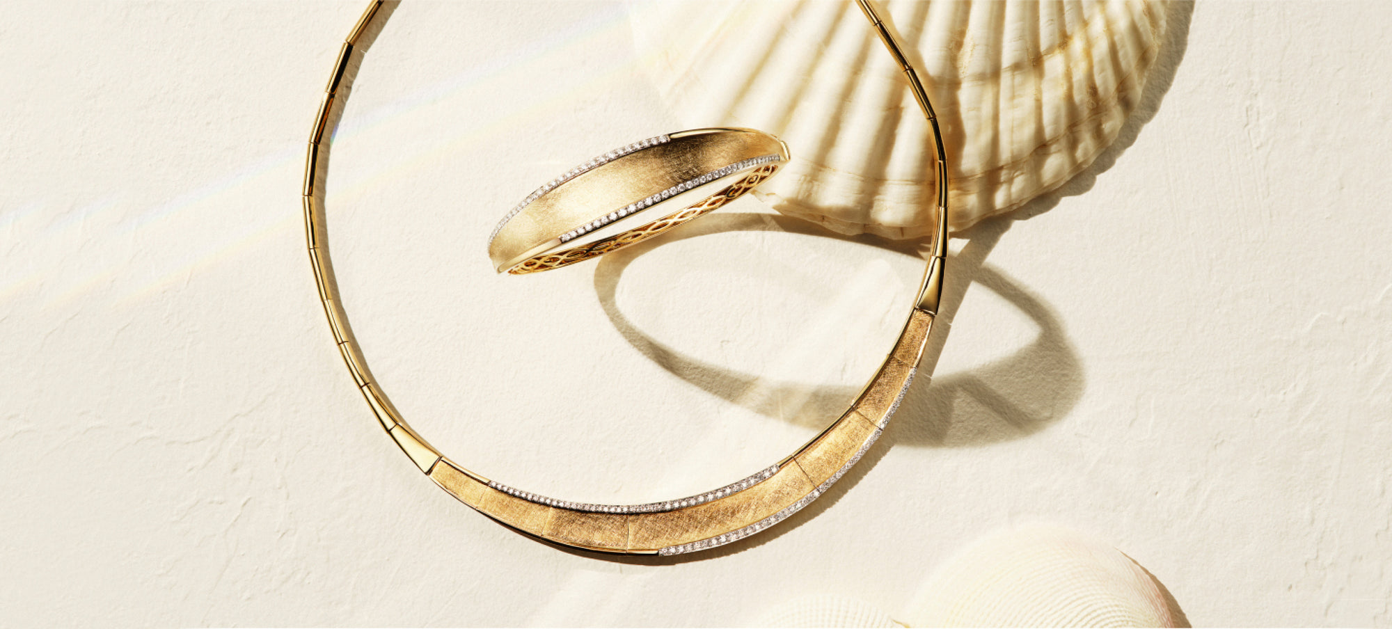 A gold necklace and matching bangle bracelet with textured surfaces and subtle diamond accents are arranged on a light sandy background with seashells.