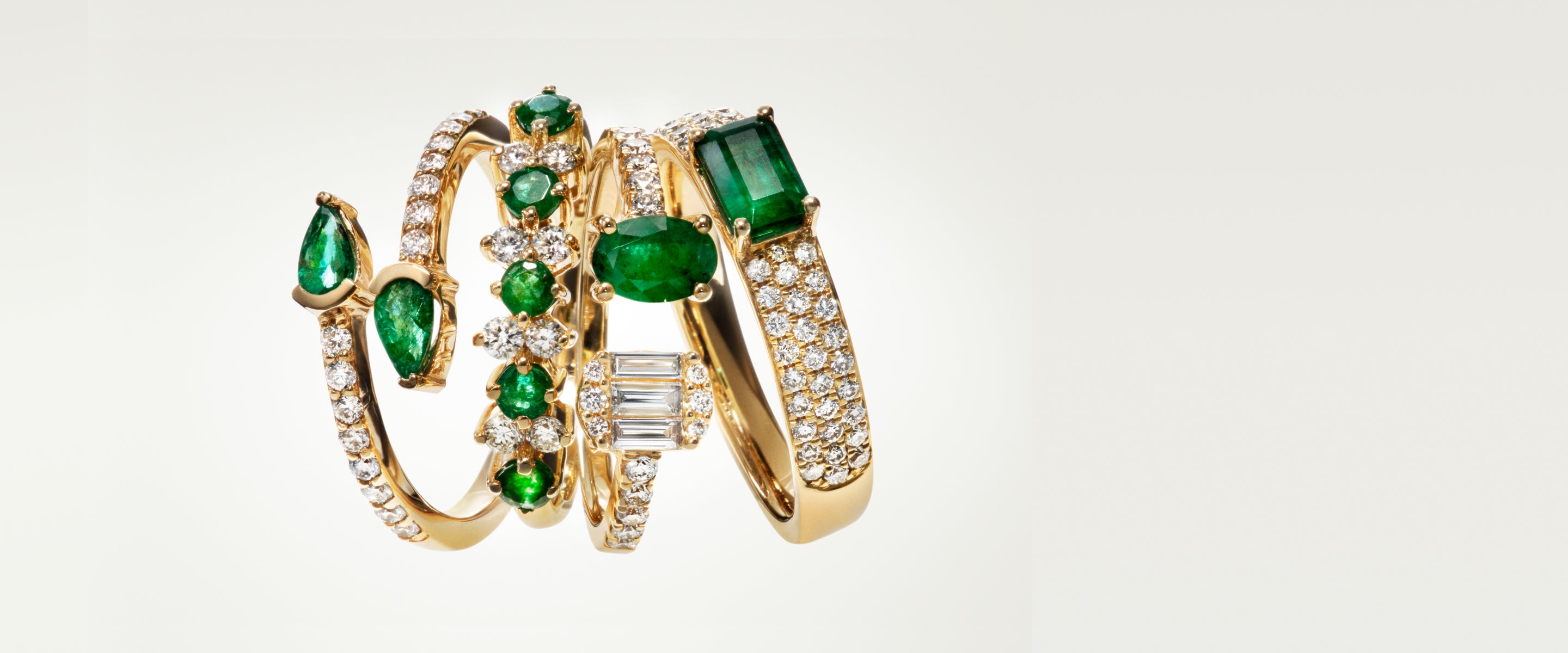 Five gold rings adorned with sparkling diamonds and vibrant green emeralds, displayed upright against a plain, light background. Each ring features a unique design and arrangement of gemstones.