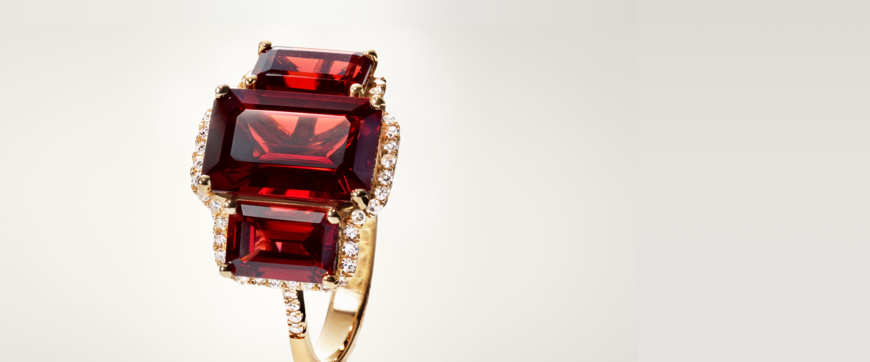 A gold ring featuring three large, rectangular red gemstones surrounded by small, sparkling diamonds is displayed against a light, neutral background.