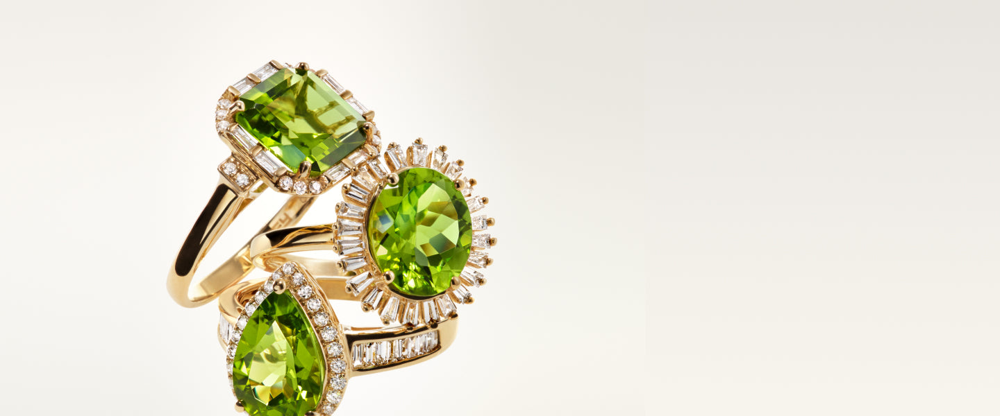 Three gold rings, each featuring a sparkling green gemstone surrounded by small diamonds, are stacked together against a plain, light background.