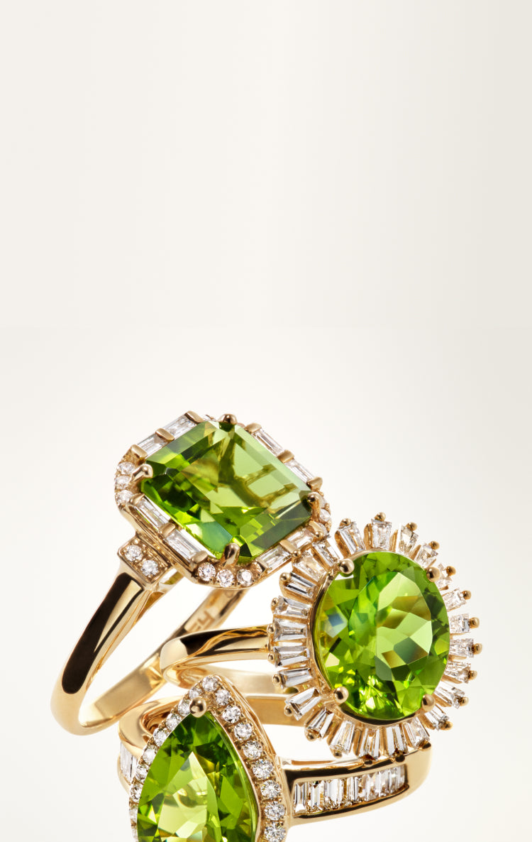 Three gold rings with vibrant green gemstones, each surrounded by sparkling clear diamonds, are elegantly stacked against a plain, light background.