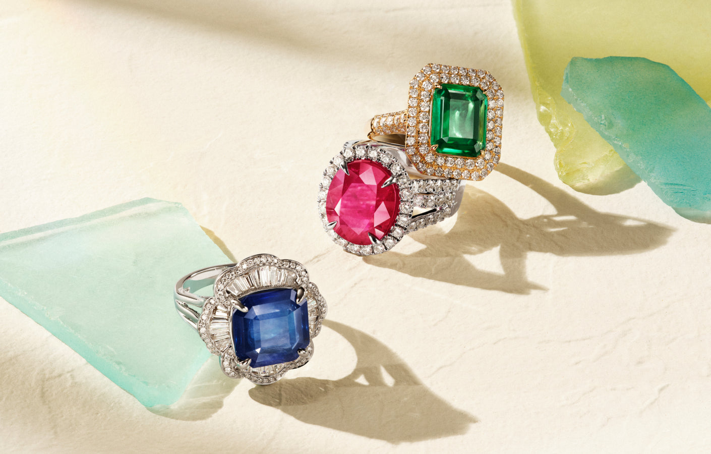 Three ornate rings with large, colorful gemstones—a blue sapphire, a pink ruby, and a green emerald—are displayed on a light surface with pastel sea glass pieces nearby. Each ring is surrounded by sparkling diamonds.