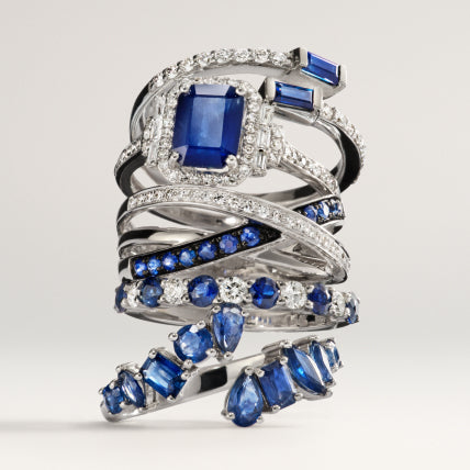 A stacked arrangement of silver rings featuring blue sapphires and white diamonds in various shapes and settings, displayed against a plain, light background.