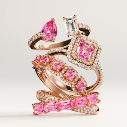 A stack of rose gold rings adorned with pink sapphires and white diamonds, featuring a variety of cuts and settings, displayed against a neutral background.