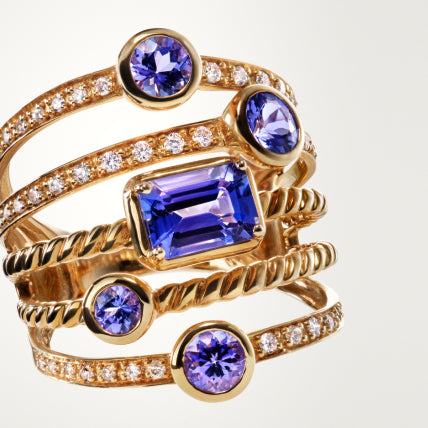 A gold ring featuring multiple bands adorned with small diamonds and five purple gemstones of varying shapes and sizes, including a large rectangular stone at the center.