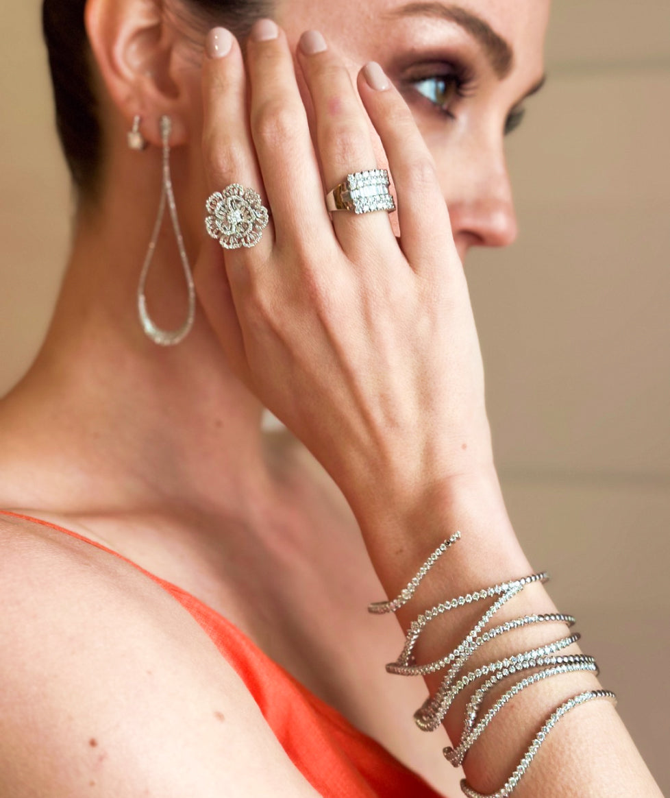 A woman in an orange dress showcases diamond jewelry, including a floral ring, stacked rings, a drop earring, and multiple coiled bracelets. Her hand is held near her face, highlighting the sparkling accessories.