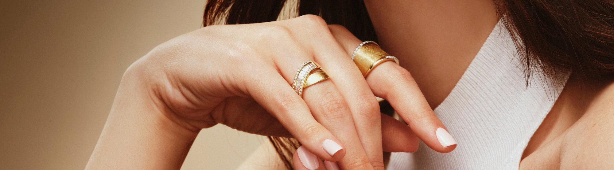 A close-up of a womans hand with light pink manicured nails, wearing two large gold rings—one plain and one with a row of diamonds—against a neutral background. She is dressed in a white, sleeveless top.