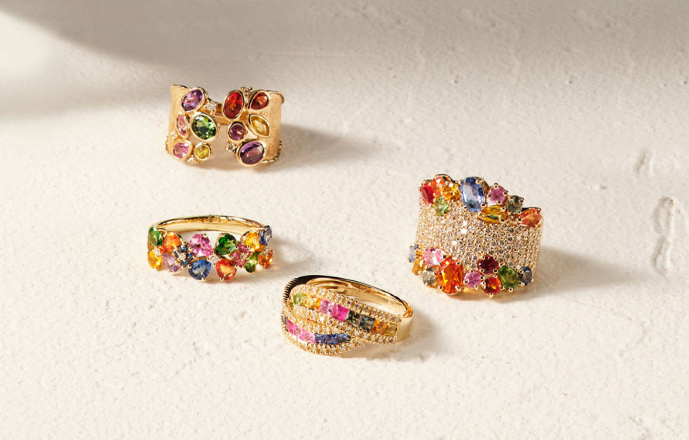Four gold rings are displayed on a textured white surface. Each ring is adorned with colorful gemstones in various shapes, including oval and round, arranged in intricate patterns.