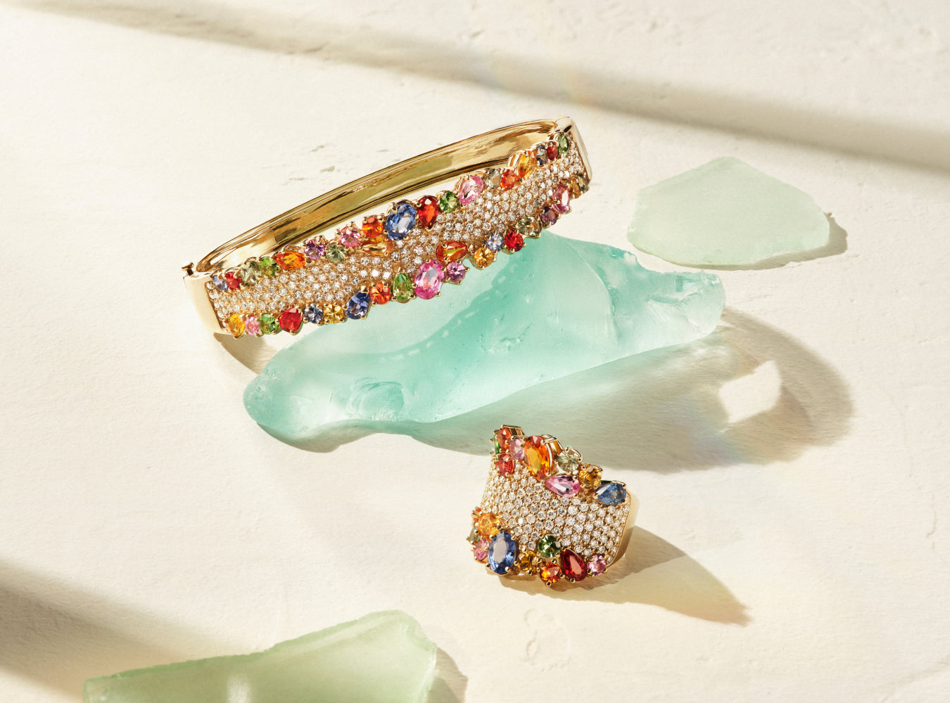 A gold bracelet and ring adorned with colorful gemstones and small crystals are displayed on light green glass pieces against a sunlit, off-white background.