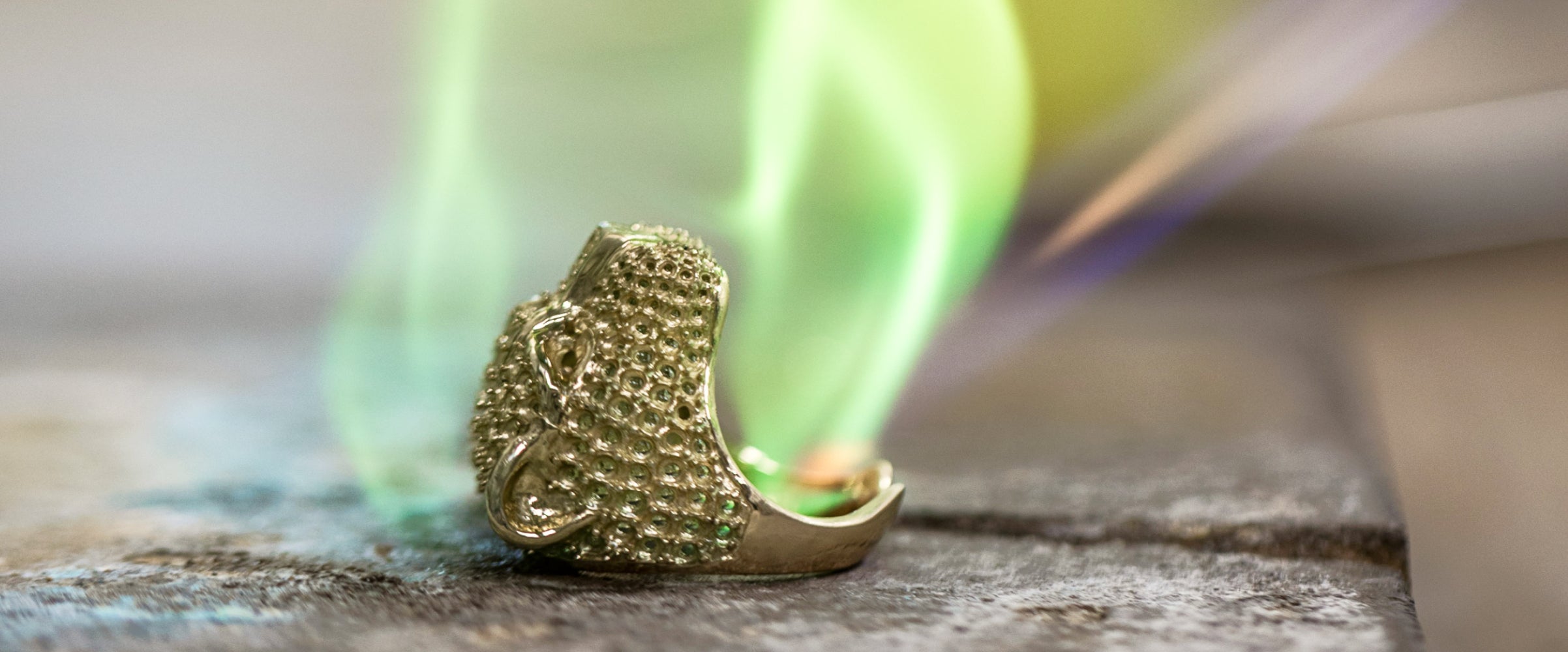 A close-up of a gold ring being crafted, with green flames or a torch flame nearby, set against a blurred background, highlighting the intricate texture and the jewelry-making process.