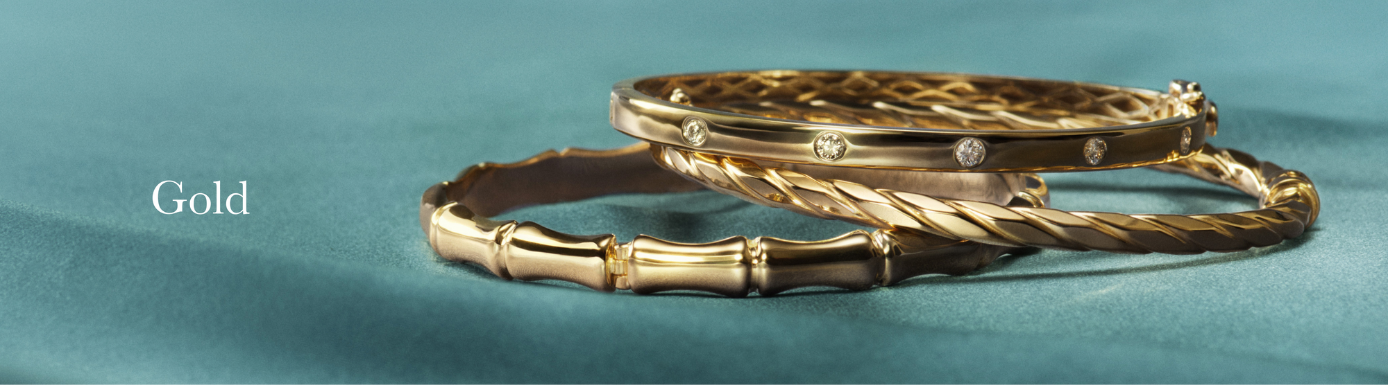 Three elegant gold bangles, one set with diamonds, rest on a soft teal fabric surface. The word Gold appears to the left in white text.