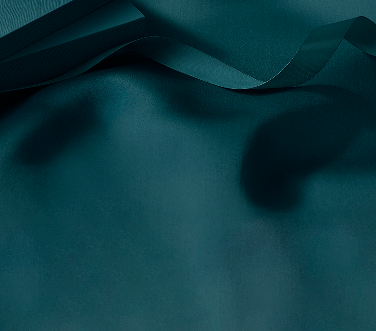A dark teal satin ribbon stretches across a matching smooth fabric surface, with the edge of a box visible in the top left corner. The scene is softly lit and elegant.