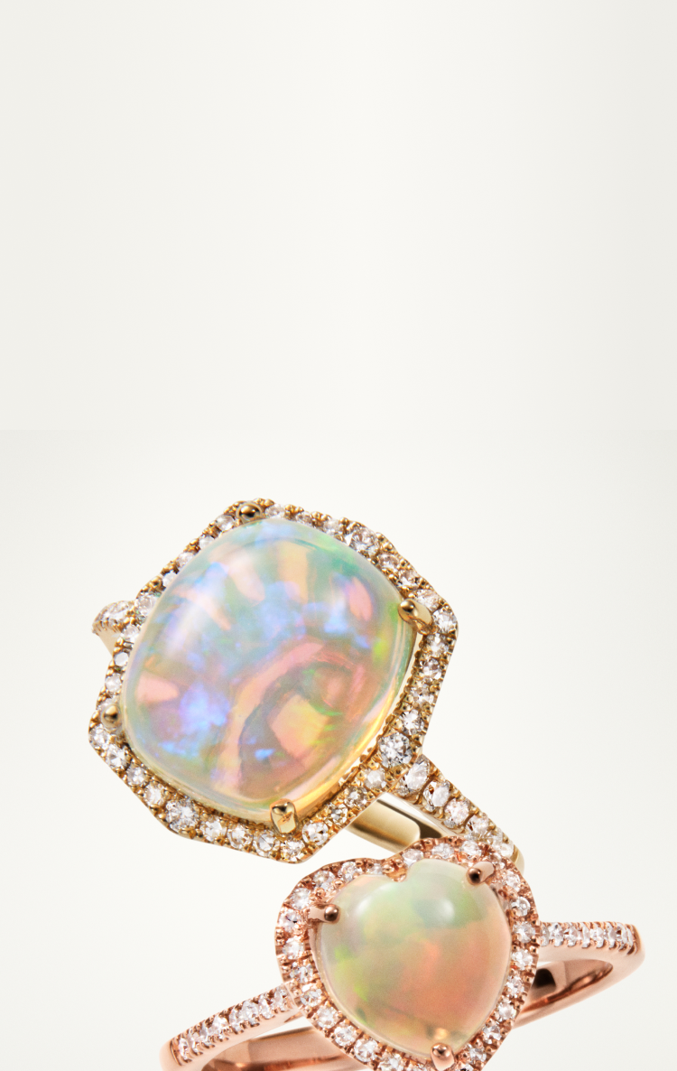 Two elegant rings with iridescent opal gemstones, each surrounded by a halo of small sparkling diamonds, are displayed on a light background. One ring is square-shaped and the other is heart-shaped.