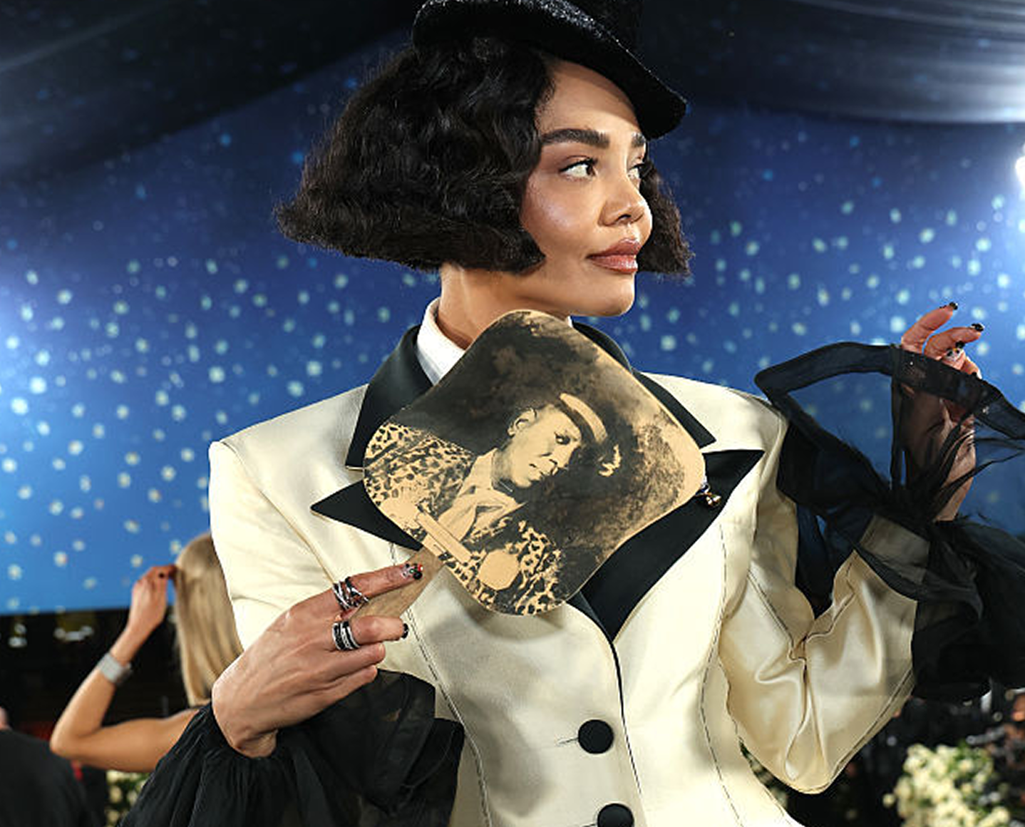 A person in a white and black suit with dramatic sleeves holds a fan featuring a vintage photo. They have styled short hair, wear multiple rings, and stand against a starry blue background.
